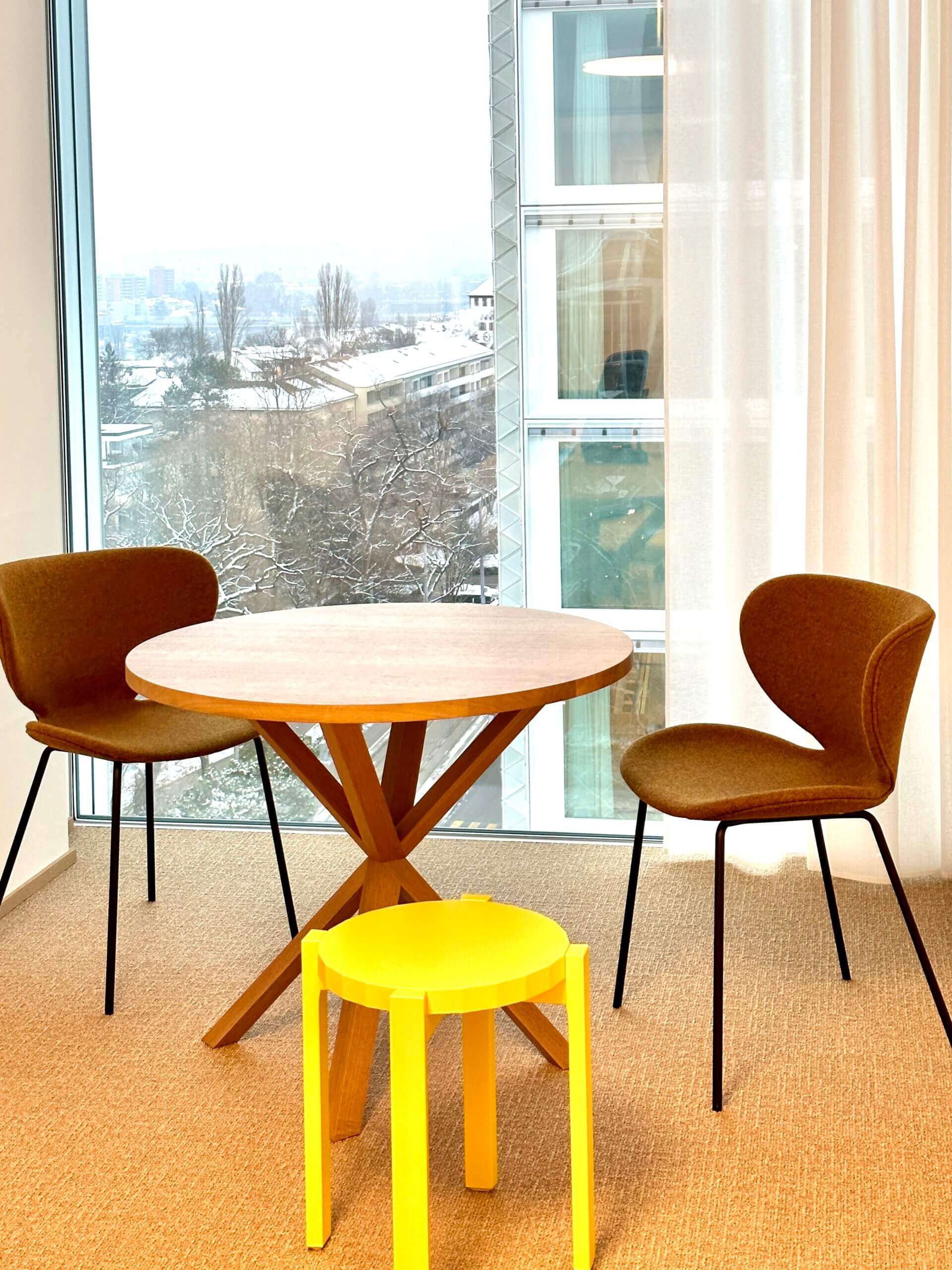 Ein helles, modern eingerichtetes Zimmer mit einem handgefertigten runden Holztisch und zwei stilvollen Stühlen, einem aus dunklem Samt und einem aus lebhaftem Gelb. Der Tisch besticht durch ein einzigartiges, sternförmiges Beindesign, das Kunsthandwerk und Funktionalität verkörpert. Im Hintergrund ist ein großes Fenster mit Blick auf winterliche Bäume und eine urbane Landschaft, was die Natürlichkeit und Nachhaltigkeit der Möbel unterstreicht. Ideal für zeitgenössische Innenarchitektur, die Wert auf handgemachte Möbel legt.