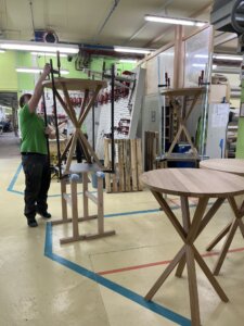 Ein Schreiner montiert sorgfältig einen Designer-Holztisch in einer gut ausgestatteten Werkstatt, umgeben von Werkzeugen und weiteren Holzelementen, die auf die qualitätsbewusste Handarbeit hinweisen.