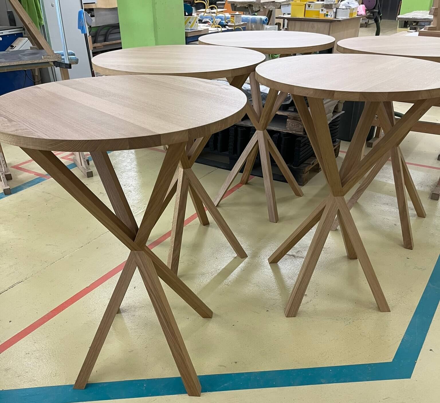 Reihe von fertiggestellten Designer-Holztischen mit charakteristischen Kreuzbein-Gestellen in einer Schreinerei, bereit für den Verkauf oder die Auslieferung.