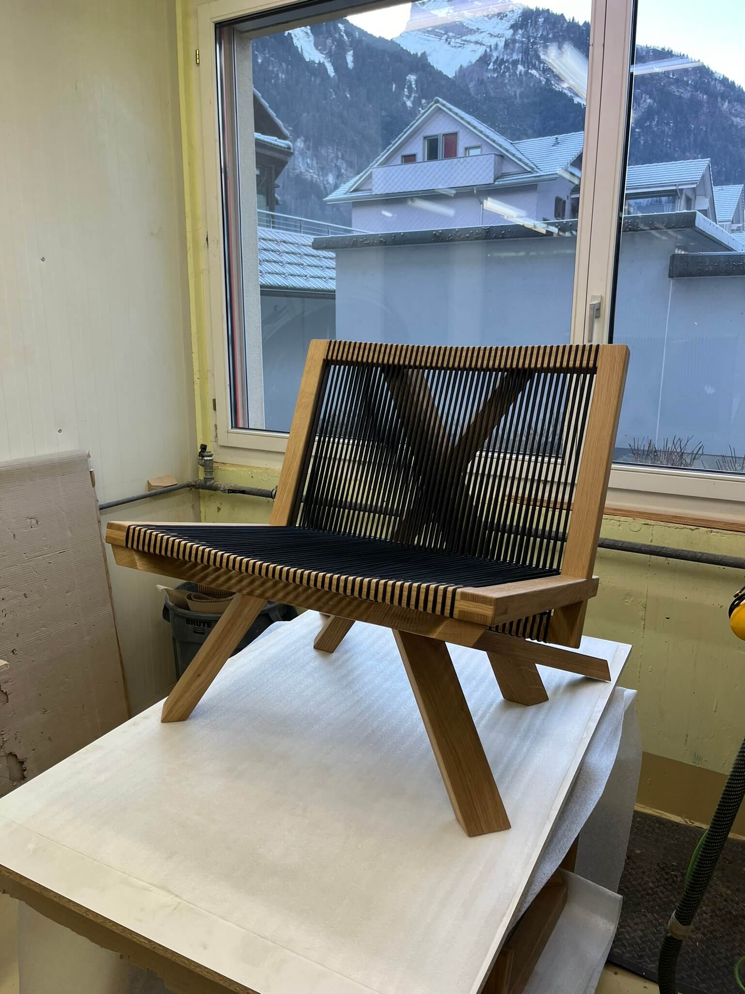 Ein neu fertiggestellter Designer-Holzsessel mit einem eleganten schwarz geflochtenen Gurtband als Sitzfläche und Rückenlehne, präsentiert vor einem Fenster mit Blick auf verschneite Berge, in einer hellen Schreinerwerkstatt.