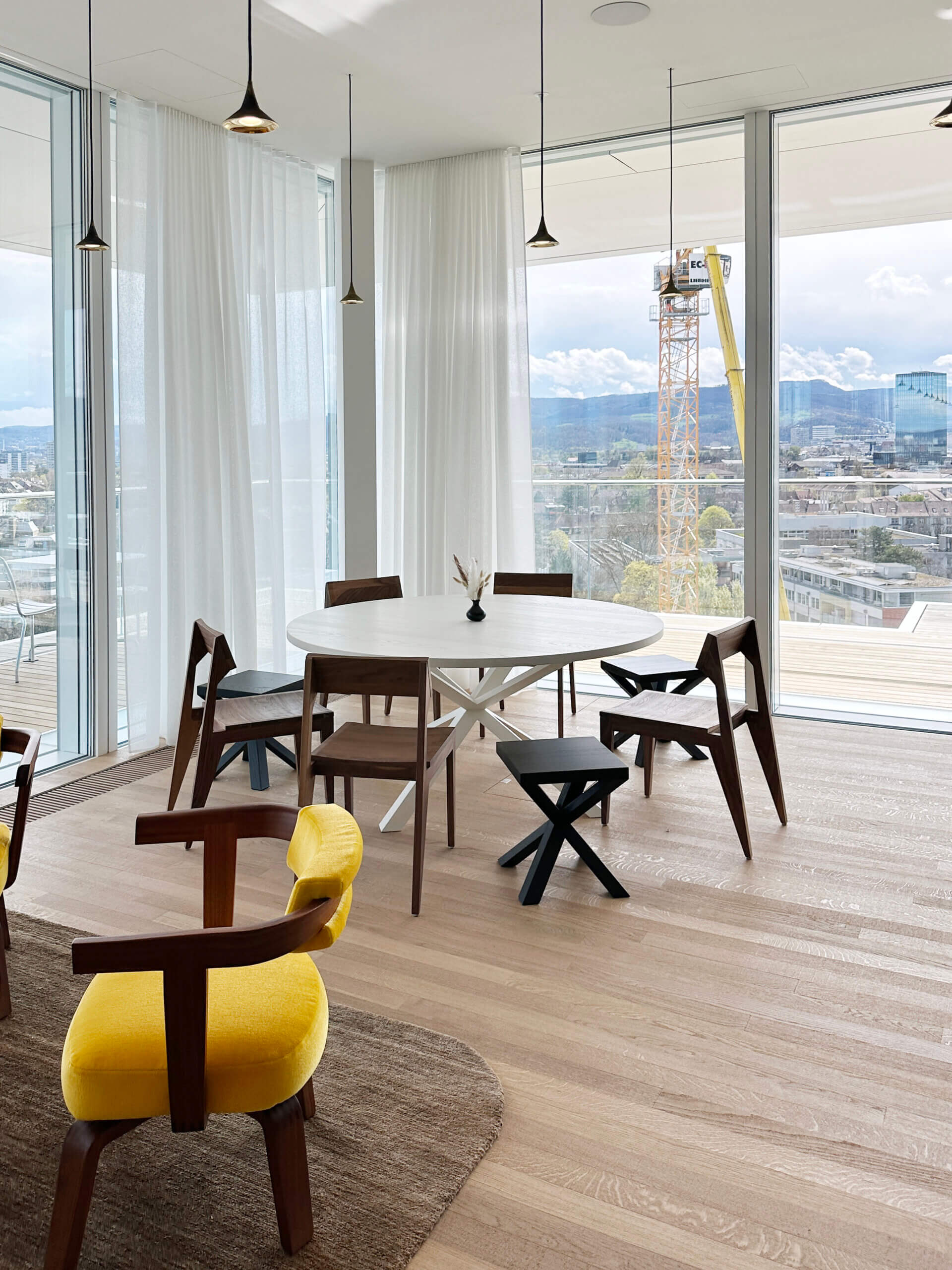 Hochwertige Möbel aus Holz in einem lichtdurchfluteten Raum mit bodentiefen Fenstern, die einen weitreichenden Blick auf die Stadt und das umliegende Bergpanorama bieten. Im Fokus stehen ein ovaler Tisch und umgebende Stühle mit einer auffälligen gelben Polsterung.