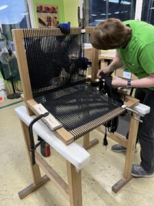 Handwerker in einer Werkstatt beim kunstvollen Flechten eines Sitzbezugs aus schwarzem Gewebe für einen handgefertigten Holzsessel, wobei Präzision und Sorgfalt im Fertigungsprozess betont werden.